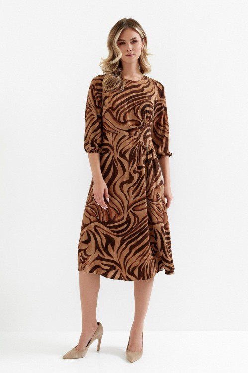 Na modelce sukienka w ciepłych odcieniach beżu i brązu, o długości midi, rozkloszowaną, z talią podkreśloną odcięciem oraz efektownym marszczeniem, z rękawami o długości 7/8 i okrągłym dekoltem