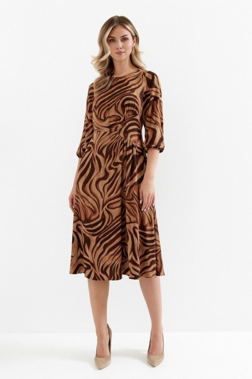 Na modelce sukienka w ciepłych odcieniach beżu i brązu, o długości midi, rozkloszowaną, z talią podkreśloną odcięciem oraz efektownym marszczeniem, z rękawami o długości 7/8 i okrągłym dekoltem