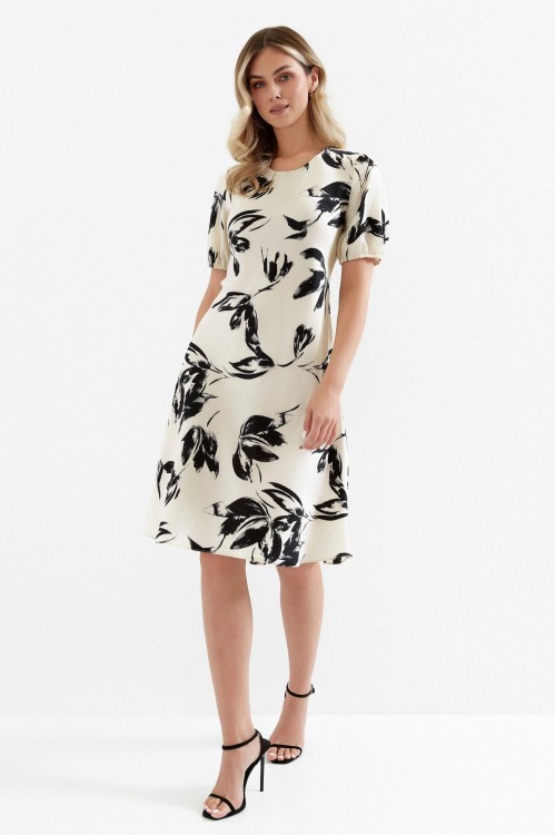 Na modelce sukienka w kolorze ecru w czarny roślinny wzór, o długości za kolano, z okrągłym dekoltem, krótki rękaw zakończony gumką