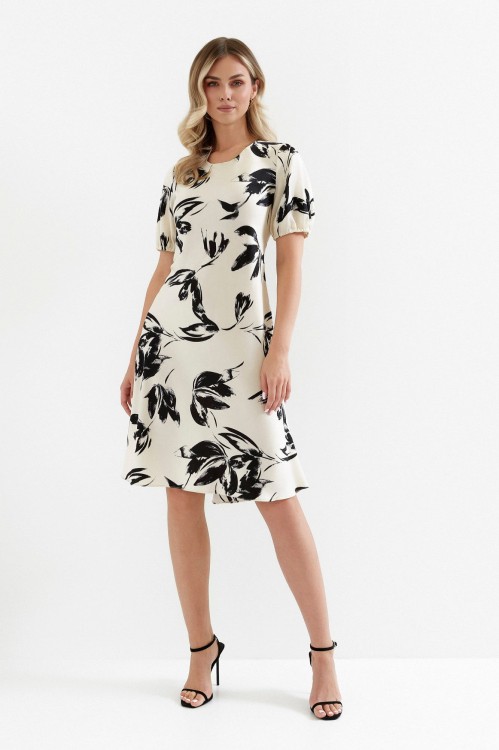Na modelce sukienka w kolorze ecru w czarny roślinny wzór, o długości za kolano, z okrągłym dekoltem, krótki rękaw zakończony gumką