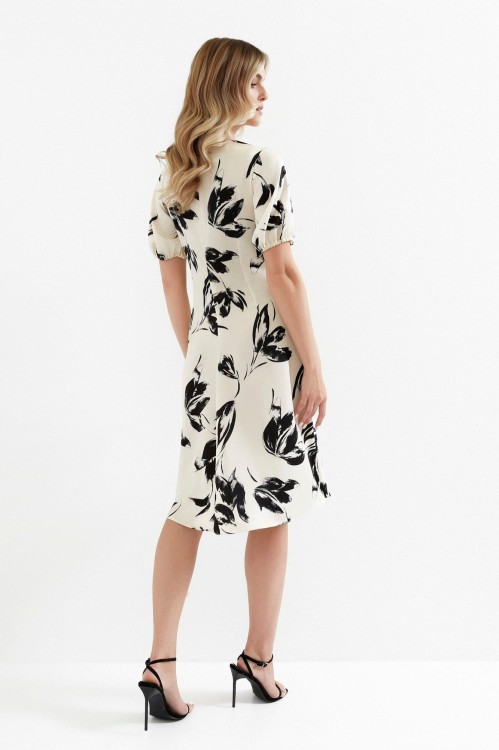 Na modelce stojącej tyłem sukienka w kolorze ecru w czarny roślinny wzór, o długości za kolano,  krótki rękaw zakończony gumką, zapinana na kryty suwak