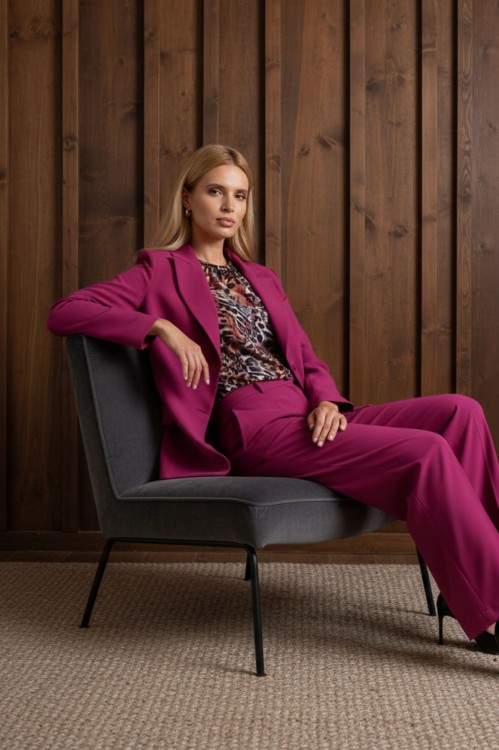 Na modelce siedzącej na krześle, krótki żakiet z wyłogami, w kolorze fuksjowym, zapinany na dwa guziki.  Do żakietu dopasowane kolorystycznie szerokie spodnie w kant