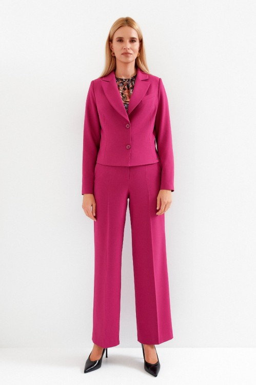 Na modelce stojącej przodem krótki żakiet z wyłogami, w kolorze fuksjowym, zapinany na dwa guziki. Na przodzie po lewej stronie imitacja kieszonki oraz długie modelujące zaszewki. Do żakietu dopasowane kolorystycznie szerokie spodnie w kant