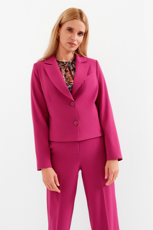 Na modelce stojącej przodem krótki żakiet z wyłogami, w kolorze fuksjowym, zapinany na dwa guziki. Na przodzie po lewej stronie imitacja kieszonki oraz długie modelujące zaszewki. Do żakietu dopasowane kolorystycznie szerokie spodnie w kant