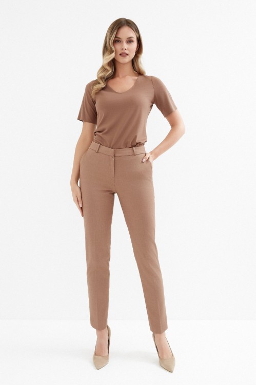 Na modelce stojącej przodem, klasyczne zwężane spodnie w kant, w kolorze ciepłego beżu, z kieszeniami, zapinane na kryty suwak, posiadającymi wszyty pasek ze szlufkami
