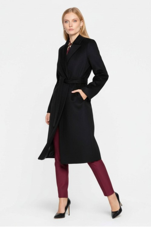 Na modelce stojącej przodem płaszcz w kolorze czarnym, z klasycznym kołnierzem, zapinany na guziki, dwurzędowy, przewiązany paskiem materiałowym, z wpuszczanymi kieszeniami o długości za kolano