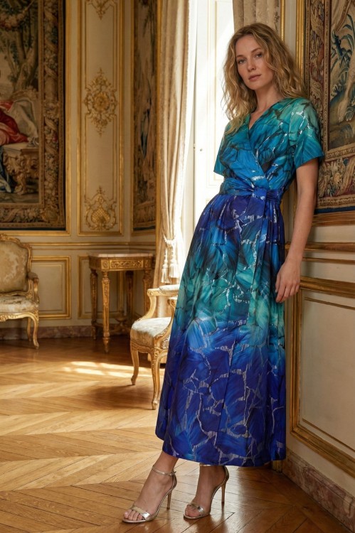 Na modelce stojącej przodem, długa sukienka w odcieniach turkusu i szafiru z metaliczną nicią w kolorze srebrnym. Sukienka ma kopertowy dekolt, krótki rękaw,  w talii przewiązana szeroką szarfą w kolorze sukienki, fason lekko rozkloszowany