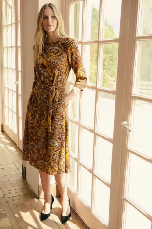 Na modelce stojącej przodem, sukienka midi w brązowy wzór paisley , z odcięciem poniżej pasa oraz falbaną, okrągły dekolt, rękaw 3/4