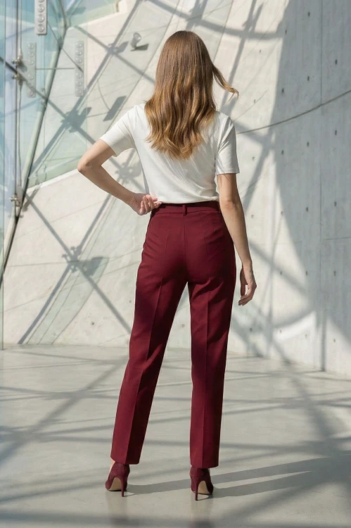 Na modelce stojącej tyłem klasyczne spodnie w kant, w kolorze bordowym, z wszywanym paskiem ze szlufkami i zaszewkami modelującymi