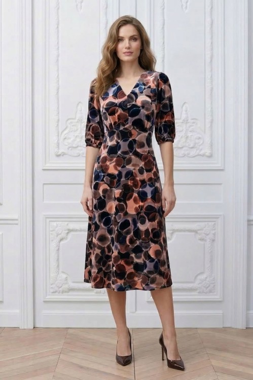 Na modelce stojącej przodem, sukienka w różnokolorowy wzór, o długości za kolana, lekko rozkloszowana, z odcięciem pod biustem w kształcie trójkąta, z lekkim przymarszczeniem, dekolt w literkę v, rękaw o długości 3/4 zakończony gumką.