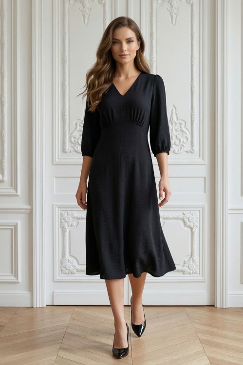 Na modelce stojącej przodem, sukienka w kolorze czarnym, o długości za kolana, lekko rozkloszowana, z odcięciem pod biustem w kształcie trójkąta, z lekkim przymarszczeniem, dekolt w literkę v, rękaw o długości 3/4 zakończony gumką.