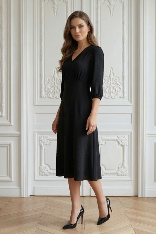Na modelce stojącej przodem, sukienka w kolorze czarnym, o długości za kolana, lekko rozkloszowana, z odcięciem pod biustem w kształcie trójkąta, z lekkim przymarszczeniem, dekolt w literkę v, rękaw o długości 3/4 zakończony gumką.