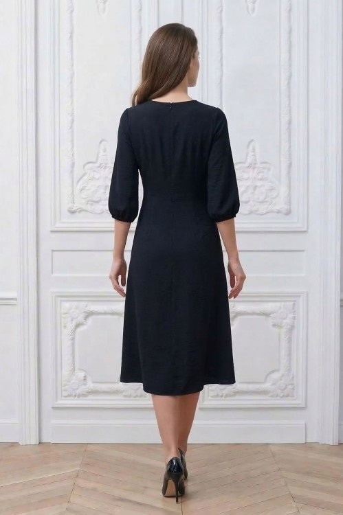 Na modelce stojącej tyłem, sukienka w kolorze czarnym, o długości za kolana, lekko rozkloszowana, rękaw o długości 3/4 zakończony gumką, zapinana na kryty suwak