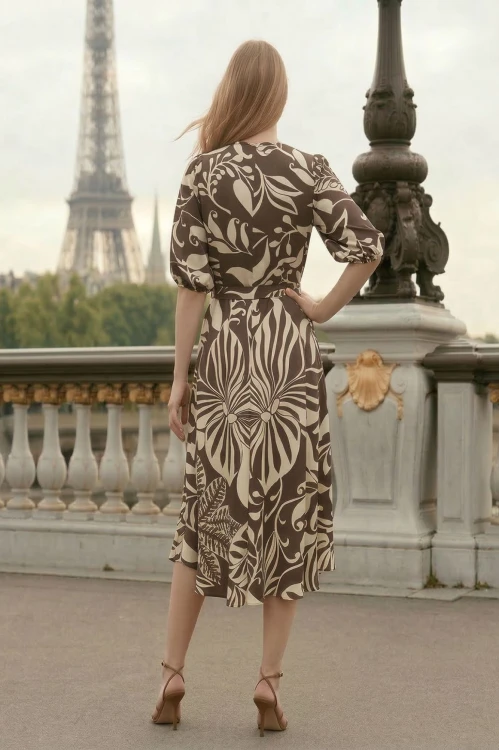 Na modelce  stojącej tyłem elegancka sukienka o długości midi, w kolorze brązowym w ecru roślinny motyw, sukienka jest przewiązywana w talii, rękawy o długości do łokcia, zapinana na plecach na długi, kryty suwak