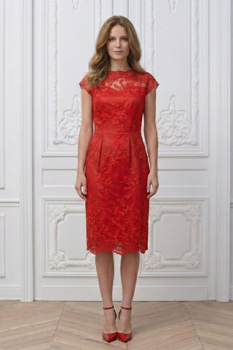 Na modelce stojącej przodem czerwona sukienka z koronki, o długości za kolana, transparentny koronkowy karczek z łódkowym dekoltem podkreśla linię ramion. Poniżej odcięcia w talii z przodu  znajdują się cztery zaszewki