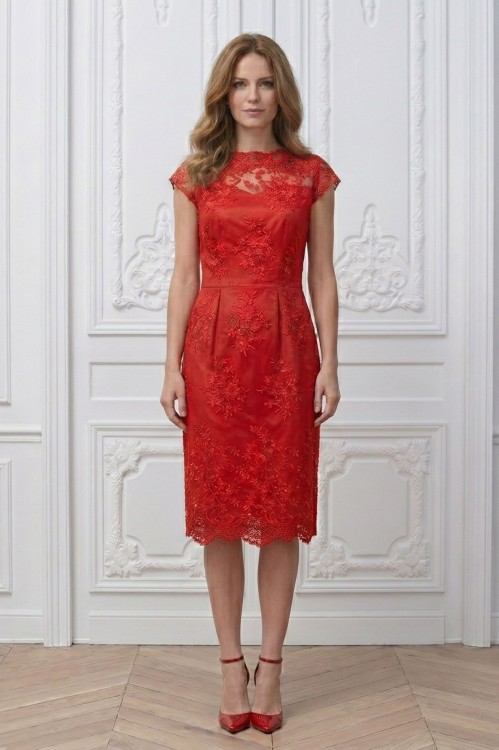 Na modelce stojącej przodem czerwona sukienka z koronki, o długości za kolana, transparentny koronkowy karczek z łódkowym dekoltem podkreśla linię ramion. Poniżej odcięcia w talii z przodu  znajdują się cztery zaszewki