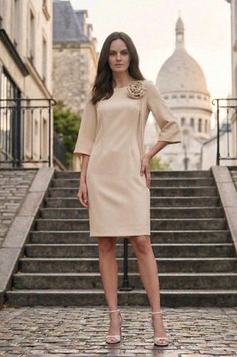 Na modelce stojącej przodem, elegancka, beżowa sukienka z przypiętą ozdobną różą. Sukienka jest o klasycznym, dopasowanym do sylwetki kroju, długości do kolan. Rękawy są lekko rozszerzane o długości 3/4