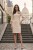 Na modelce stojącej przodem, elegancka, beżowa sukienka z przypiętą ozdobną różą. Sukienka jest o klasycznym, dopasowanym do sylwetki kroju, długości do kolan. Rękawy są lekko rozszerzane o długości 3/4