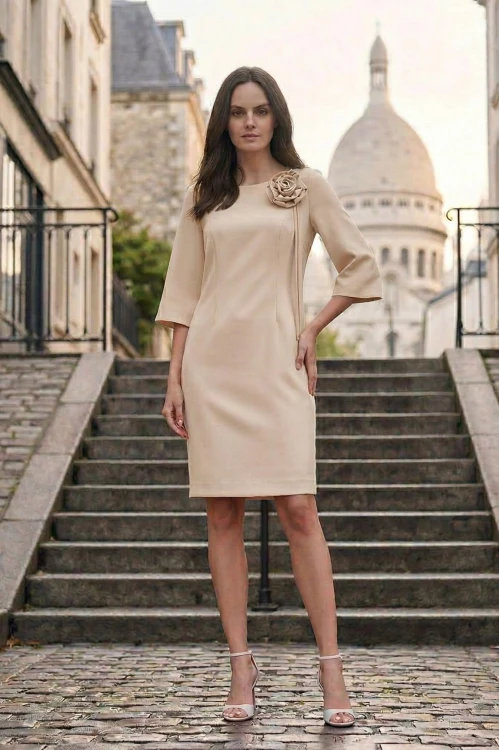 Na modelce stojącej przodem, elegancka, beżowa sukienka z przypiętą ozdobną różą. Sukienka jest o klasycznym, dopasowanym do sylwetki kroju, długości do kolan. Rękawy są lekko rozszerzane o długości 3/4
