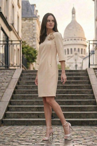 Na modelce stojącej w pozie lekko bokiem, elegancka, beżowa sukienka z przypiętą ozdobną różą. Sukienka jest o klasycznym, dopasowanym do sylwetki kroju, długości do kolan. Rękawy są lekko rozszerzane o długości 3/4