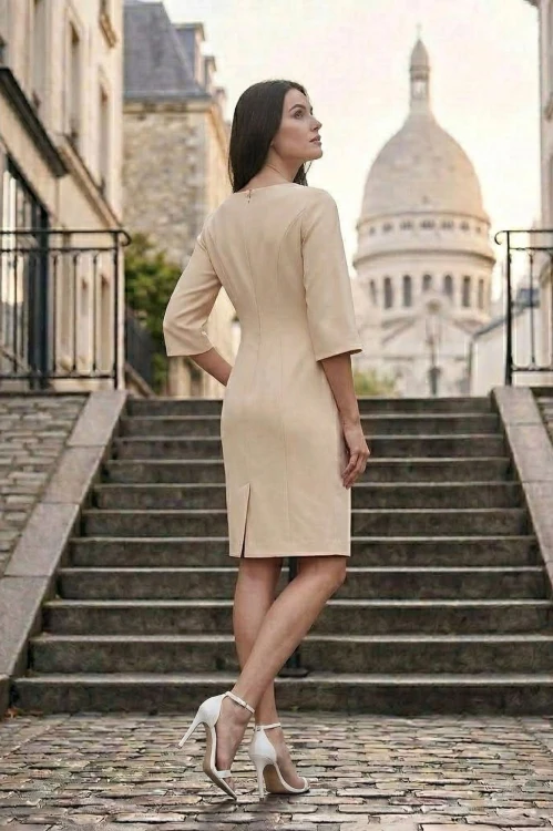 Na modelce stojącej w pozie tyłem, lekko bokiem, beżowa sukienka o d€gości do kolan. Dopasowana do sylwetki, zaszewkami modelującymi. Zapinana na kryty suwak. U dołu kryty rozporek.