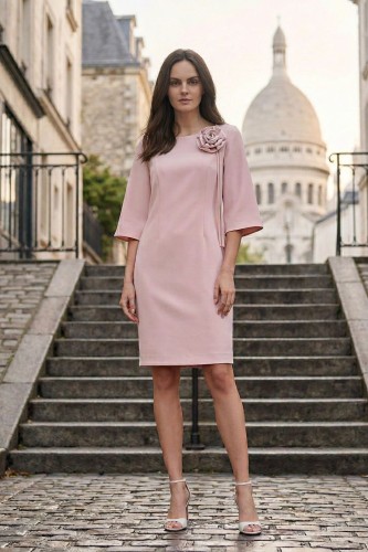 Na modelce stojącej przodem, elegancka, jasnoróżowa sukienka z przypiętą ozdobną różą. Sukienka jest o klasycznym, dopasowanym do sylwetki kroju, długości do kolan. Rękawy są lekko rozszerzane o długości 3/4