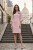 Na modelce stojącej przodem, elegancka, jasnoróżowa sukienka z przypiętą ozdobną różą. Sukienka jest o klasycznym, dopasowanym do sylwetki kroju, długości do kolan. Rękawy są lekko rozszerzane o długości 3/4