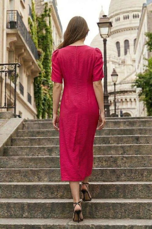 Na modelce stojącej tyłem midi rozszerzana ku dołowi sukienka, w kolorze malinowym.  Sukienka ma krótkie, bufiaste rękawy, okrągły dekolt i długi kryty suwak. Dopasowana do sylwetki, długimi zaszewkami modelującymi