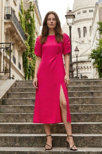 Na modelce stojącej przodem midi sukienka w odcieniu malinowym, rozszerzana ku dołowi, z długim rozcięciem eksponującym kolano. sukienka ma okrągły dekolt i krótkie, bufiaste rękawki z ozdobnym marszczeniem