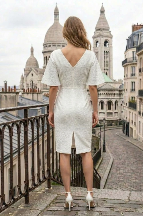 Na modelce stojącej tyłem, sukienka w kolorze białym, o długości do kolan, z rozporkiem, dekolt w literkę v, rękaw krótki rozszerzany, zapinana na długi kryty suwak, zaszewki modelujące,