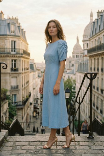 Na modelce stojącej przodem sukienka midi w odcieniu niebieskim, rozszerzana ku dołowi, z długim rozcięciem, sukienka ma okrągły dekolt i krótkie, bufiaste rękawki z ozdobnym marszczeniem