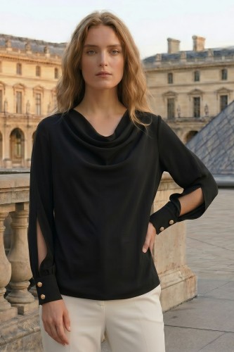 Na modelce stojącej przodem Elegancka czarna bluzka z dekoltem typu woda, długimi rękawami zakończonymi mankietami zapinanymi na dwa ozdobne guziki, powyżej mankietów efektowne rozcięcia