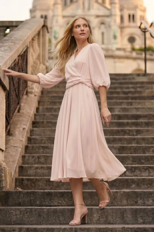 Na modelce stojącej przodem sukienka midi w kolorze pudrowego różu, dekolt kopertowy, odcięcie w pasie ozdobione efektownym drapowaniem, dół zwiewny rozkloszowany, rękawy 3/4 zakończone delikatną gumką