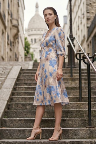 Na modelce stojącej lekko bokiem, sukienka w niebieskie kwiaty na beżowym tle, o długości za kolana, lekko rozkloszowana, z odcięciem pod biustem w kształcie trójkąta, z lekkim przymarszczeniem, dekolt w literkę v, rękaw o długości 3/4 zakończony gumką.