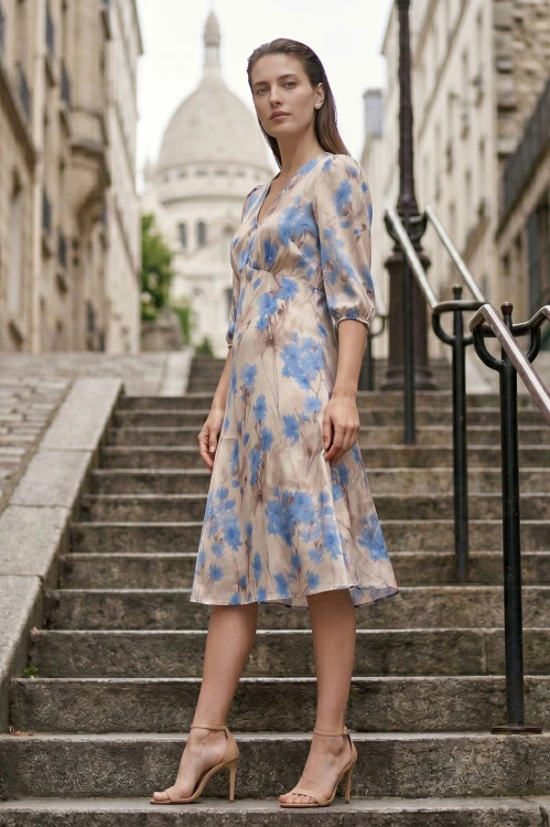 Na modelce stojącej lekko bokiem, sukienka w niebieskie kwiaty na beżowym tle, o długości za kolana, lekko rozkloszowana, z odcięciem pod biustem w kształcie trójkąta, z lekkim przymarszczeniem, dekolt w literkę v, rękaw o długości 3/4 zakończony gumką.