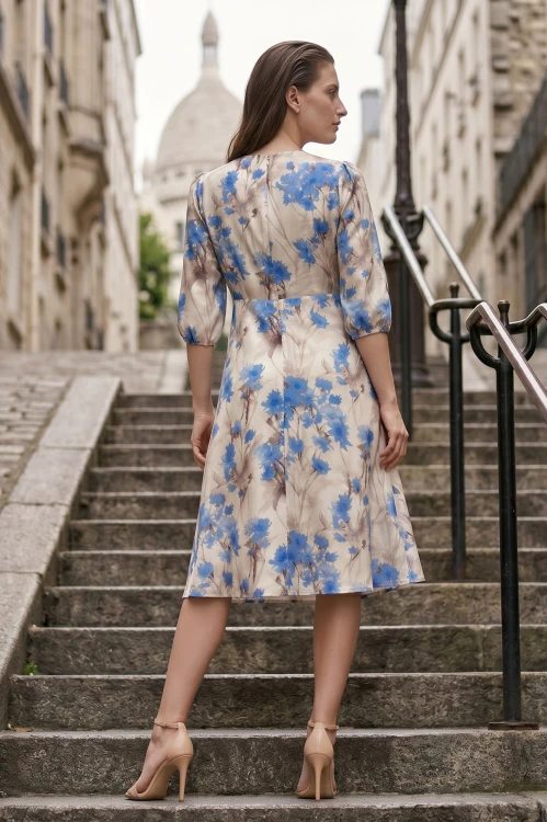 Na modelce stojącej tyłem, sukienka w  w niebieskie kwiaty na beżowym tle, o długości za kolana, lekko rozkloszowana, zapinana na kryty suwak, rękaw o długości 3/4 zakończony gumką