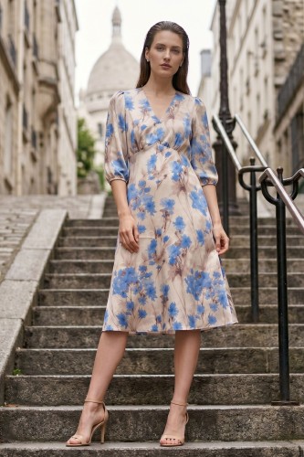 Na modelce stojącej przodem, sukienka w niebieskie kwiaty na beżowym tle, o długości za kolana, lekko rozkloszowana, z odcięciem pod biustem w kształcie trójkąta, z lekkim przymarszczeniem, dekolt w literkę v, rękaw o długości 3/4 zakończony gumką.