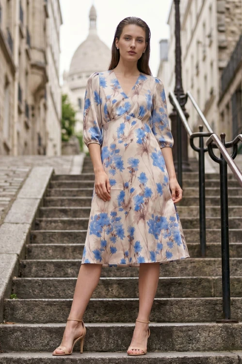 Na modelce stojącej przodem, sukienka w niebieskie kwiaty na beżowym tle, o długości za kolana, lekko rozkloszowana, z odcięciem pod biustem w kształcie trójkąta, z lekkim przymarszczeniem, dekolt w literkę v, rękaw o długości 3/4 zakończony gumką.