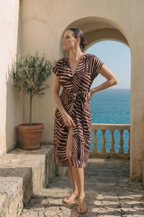 Na modelce stojącej przodem sukienka w zwierzęcy print w kolorach brązu i beżu, z kimonowym, krótkim rękawem, przewiązywana w talii materiałowym paskiem w tym samym kolorze. Dekolt w kształcie V. Długość za kolana z rozcięciem z przodu
