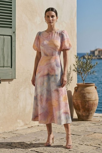 Na modelce sukienka o długości midi  rozkloszowana w pastelowych kolorach, z okrągły dekoltem, odcięciem po wyżej talii, krótkim rękawkiem zakończonym gumką, tworząc bufkę.