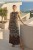 Na modelce, czarna sukienka maxi w ornamentalny kremowo-brązowy wzór, sukienka ma okrągły dekolt otoczony ozdobną plecionką, ramiona odkryte, w talii delikatna gumka z ozdobnym sznureczkiem. Na dole sukienki, po bokach wysokie rozcięcia.