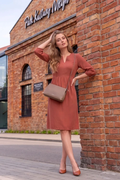 Na modelce stojącej przodem, sukienka w kolorze herbacianym, o długości do kolan. Sukienka ma odcięcie lekko powyżej talii, dekolt w v oraz wszywane rękawy długości 3/4