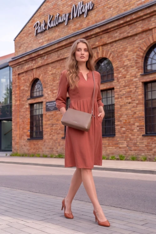 Na modelce stojącej przodem, sukienka w kolorze herbacianym, o długości do kolan. Sukienka ma odcięcie lekko powyżej talii, dekolt w v oraz wszywane rękawy długości 3/4, posiada także kieszenie