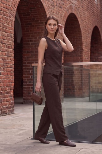 Na modelce eleganckie spodnie w kant, w kolorze brązowym. Jest to model o szerokiej nogawce z zaprasowanymi kantami. Po bokach, wpuszczane w boczne szwy, kieszenie. Do spodni dopasowana kamizelka