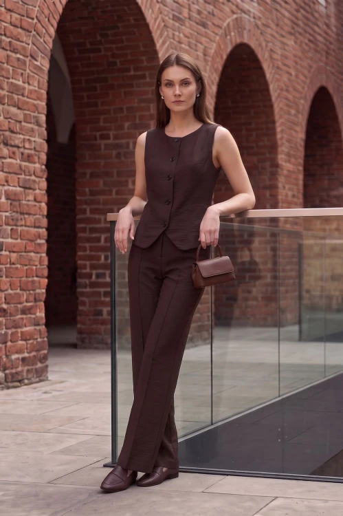 Na modelce elegancka kamizelka w brązowym kolorze, model ma okrągły dekolt, zaszewki dopasowujące do sylwetki, oraz lekkie zaokrąglenia u dołu. Do kamizelki dopasowane kolorystycznie spodnie, tworzą spójny komplet