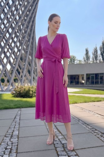 Na modelce stojącej przodem sukienka kolorze fuksji, o długości midi, rozkloszowana, z kopertowym dekoltem, 3/4 rękawami i materiałowym paskiem w talii