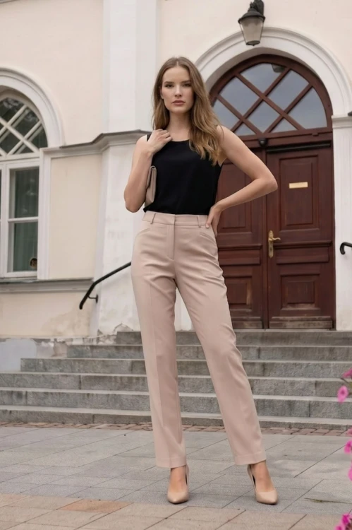 Na modelce stojącej przodem klasyczne spodnie w kant, w kolorze jasnobeżowym, zapinane z przodu na kryty suwak i haftkę, z wszywanym paskiem ze szlufkami