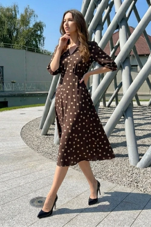 Na modelce sukienka w kolorze czekoladowym w beżowe groszki, o długości midi, lekko rozkloszowana, z odcięciem pod biustem w kształcie trójkąta, z lekkim przymarszczeniem, dekolt w literkę v, rękaw o długości 7/8 zakończony gumką.