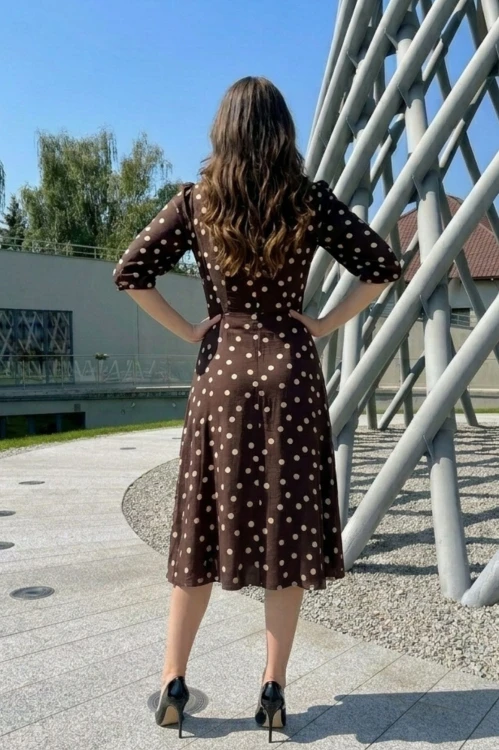 Na modelce stojącej tyłem sukienka w kolorze  czekoladowy w beżowegroszki, o długości midi, lekko rozkloszowana,  rękaw o długości 7/8 zakończony gumką.