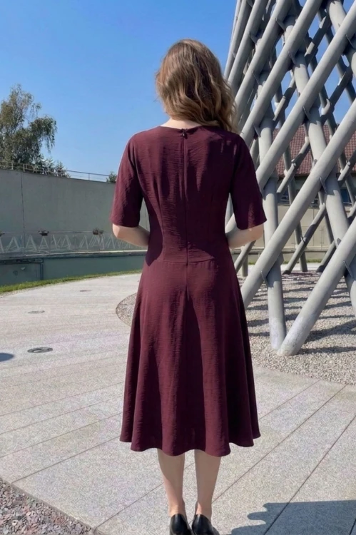 Na modelce stojącej tyłem, sukienka midi w kolorze bordowym, z obniżoną talią, rękawek kimonowy o długości do łokcia,  lekko rozkloszowany, zapinana na kryty suwak